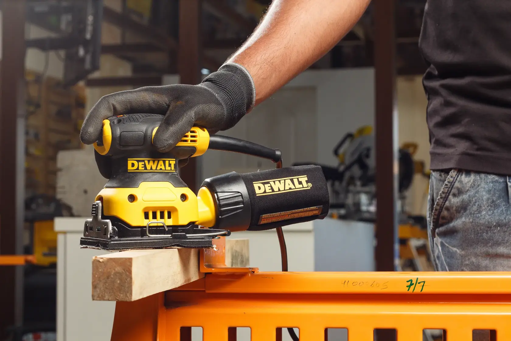 Vista de primer plano de una mano enguantada usando una lijadora eléctrica amarilla y negra DEWALT DWE6411 sobre una superficie de madera al aire libre.