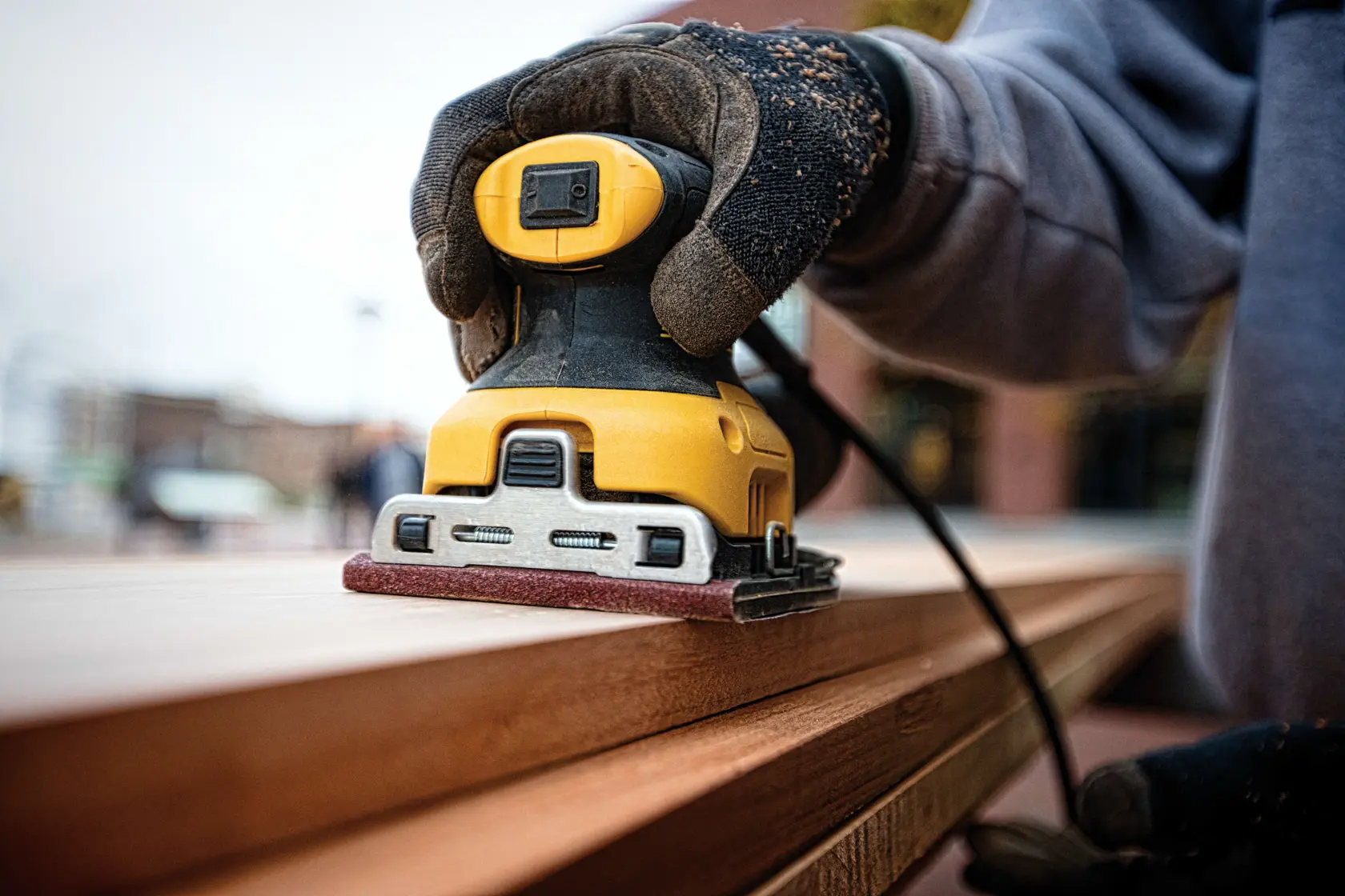 Primer plano de una persona con guantes usando una lijadora eléctrica amarilla DEWALT para suavizar la superficie de una tabla de madera.
