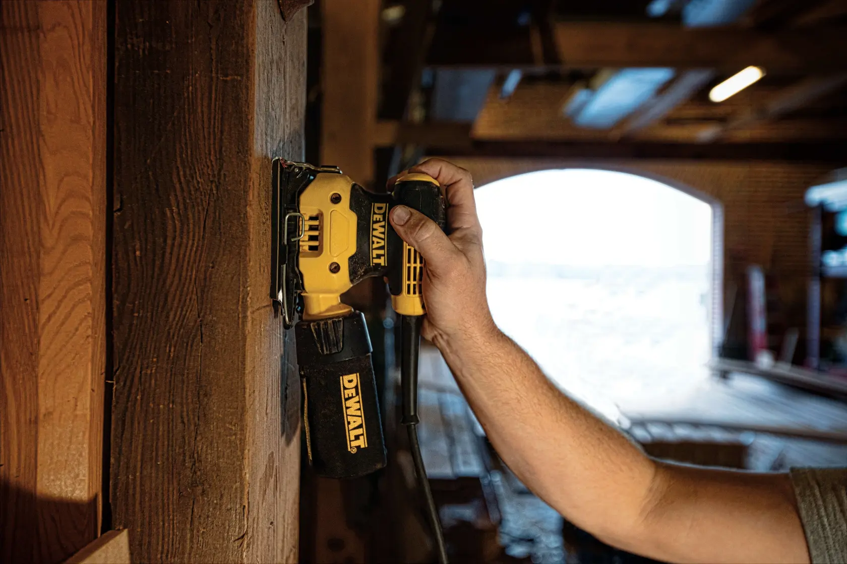 Una persona está utilizando una lijadora eléctrica DEWALT DWE6411 en una superficie de madera dentro de un taller, con la lijadora sostenida verticalmente y la bolsa para polvo acoplada.