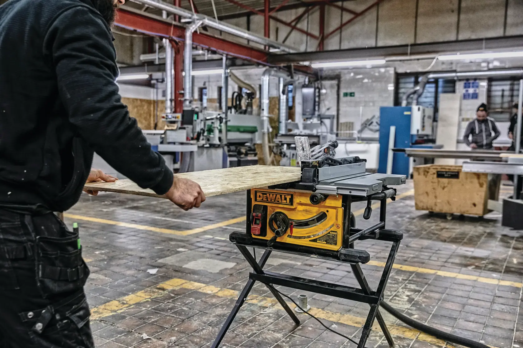 Una persona está utilizando una sierra de mesa amarilla DEWALT (modelo DWE7492SET) montada en un soporte negro para cortar una tabla de madera dentro de un taller con maquinaria industrial al fondo.