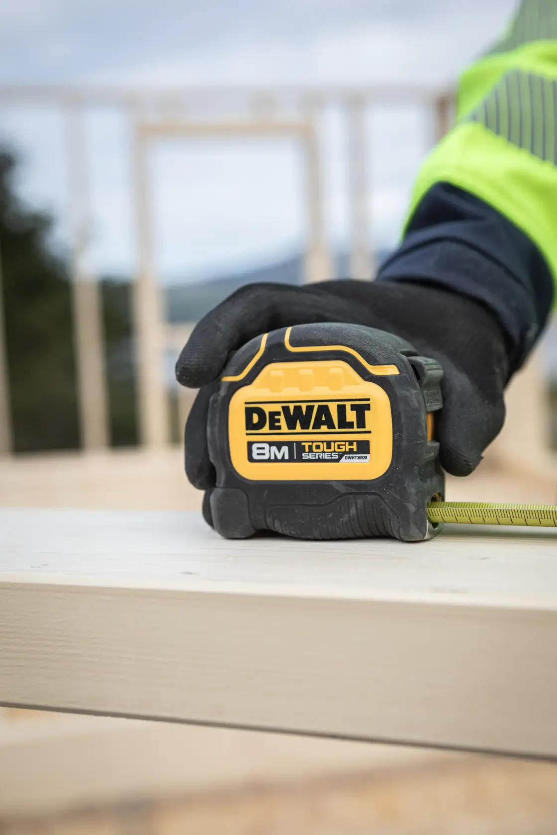 Cinta métrica DEWALT 8M Tough Series sostenida por una mano enguantada sobre tabla en construcción.