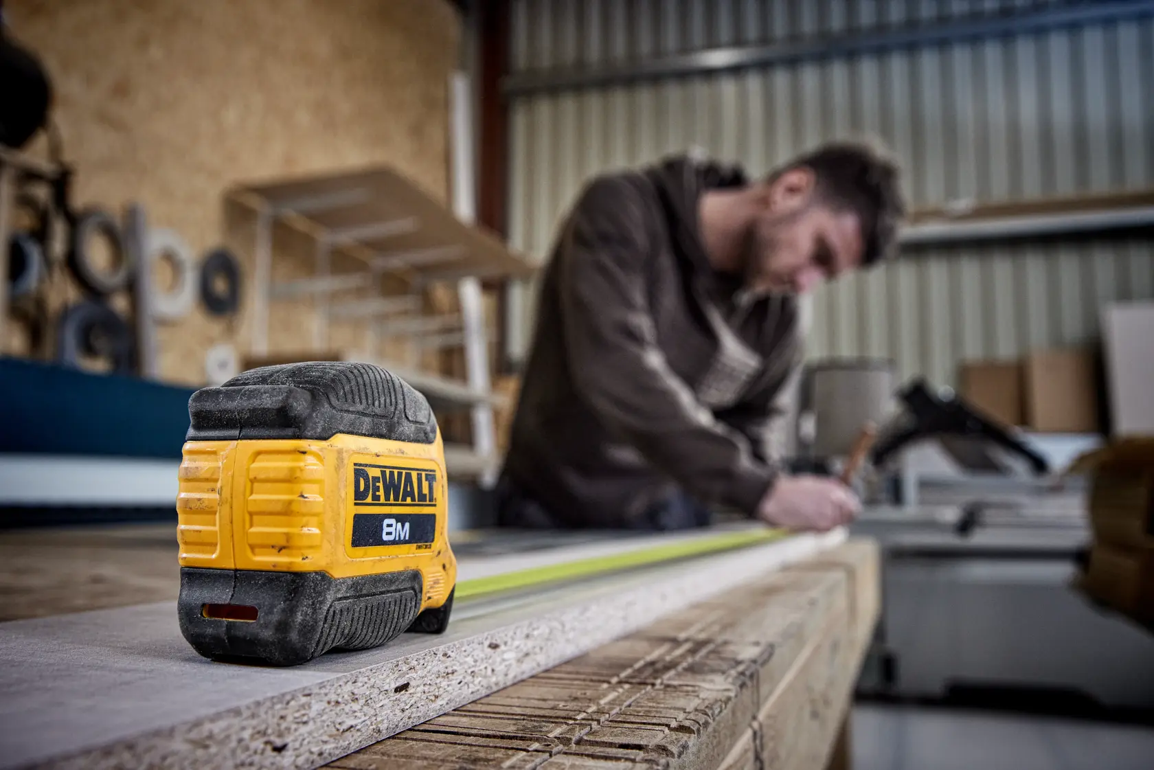 Cinta métrica DEWALT 8M sobre madera, persona midiendo al fondo en un taller.