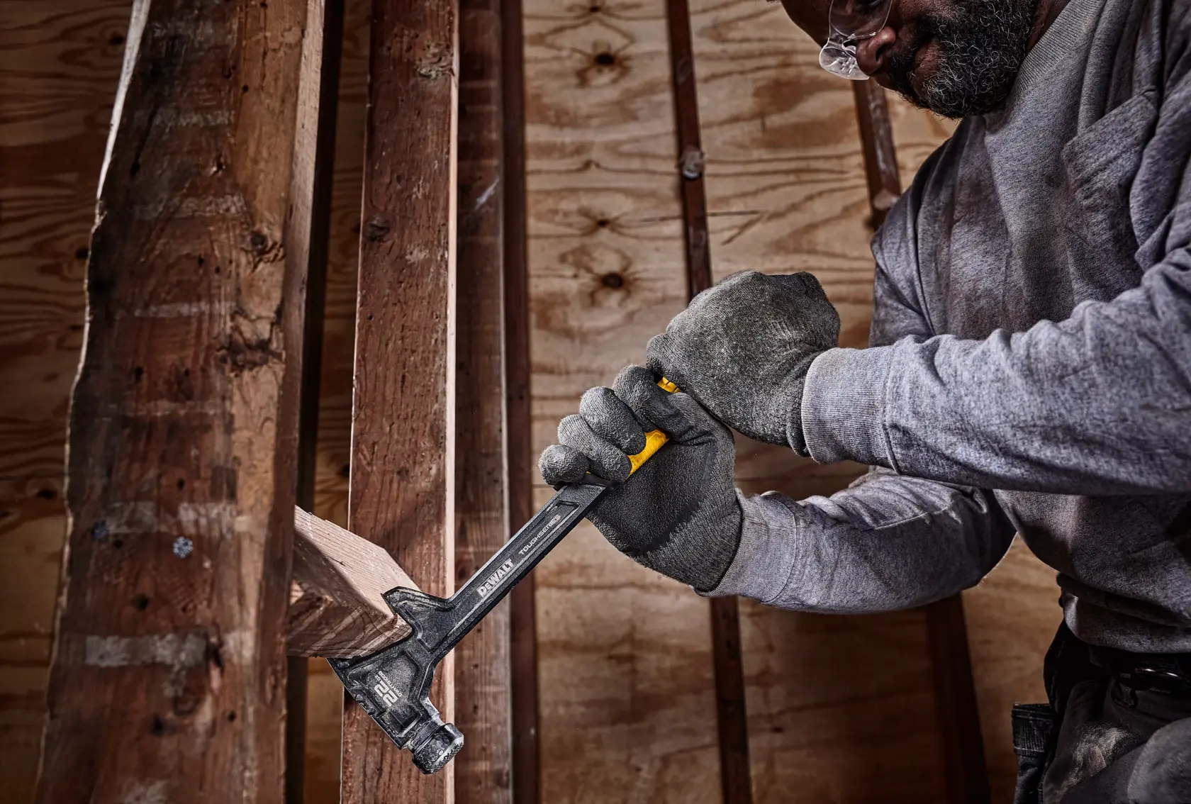 Una persona con guantes de trabajo utiliza un martillo DEWALT para quitar una tabla de madera en un entorno de construcción.