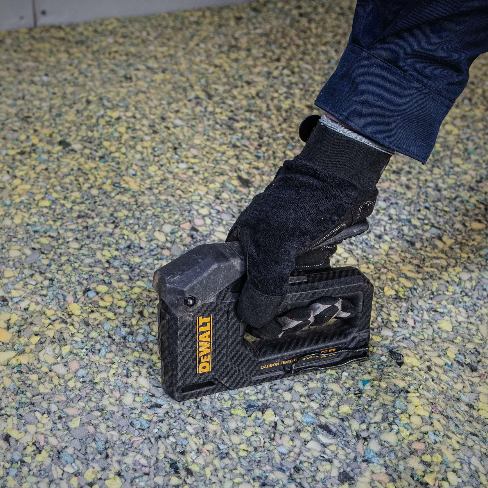 A close-up of a gloved hand holding a DEWALT carbon fiber composite hand tool, positioned on a textured surface with small colored stones.