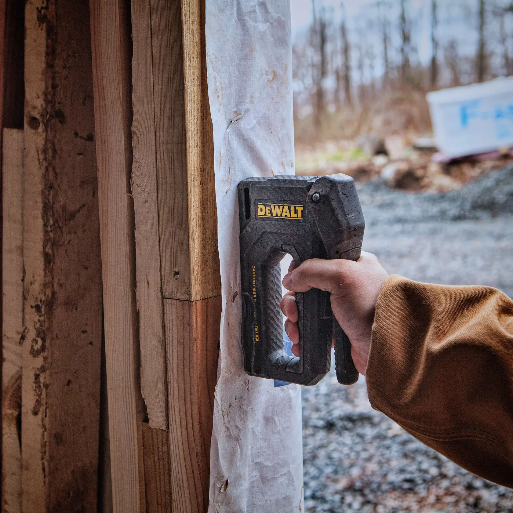 A person wearing a black work glove uses a DEWALT hand stapler to attach insulation material to a wooden wall.