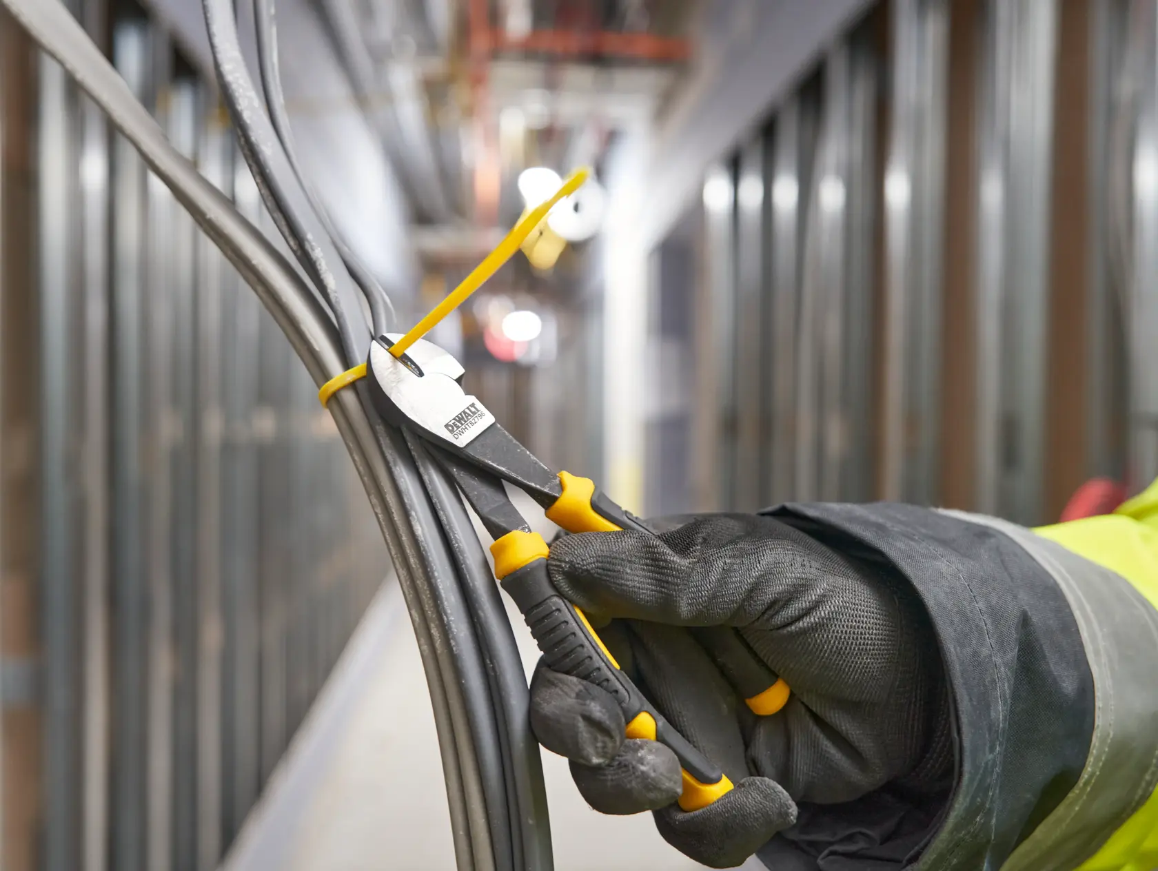 Mano con guante usa alicate DEWALT para cortar brida que sujeta varios cables eléctricos juntos en taller.