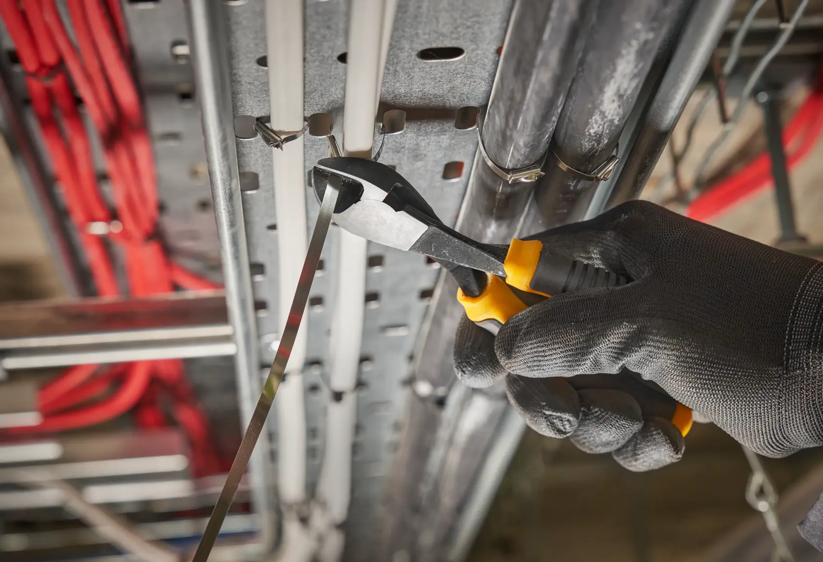 Mano enguantada usa pinzas DEWALT para cortar brida metálica en instalación eléctrica.