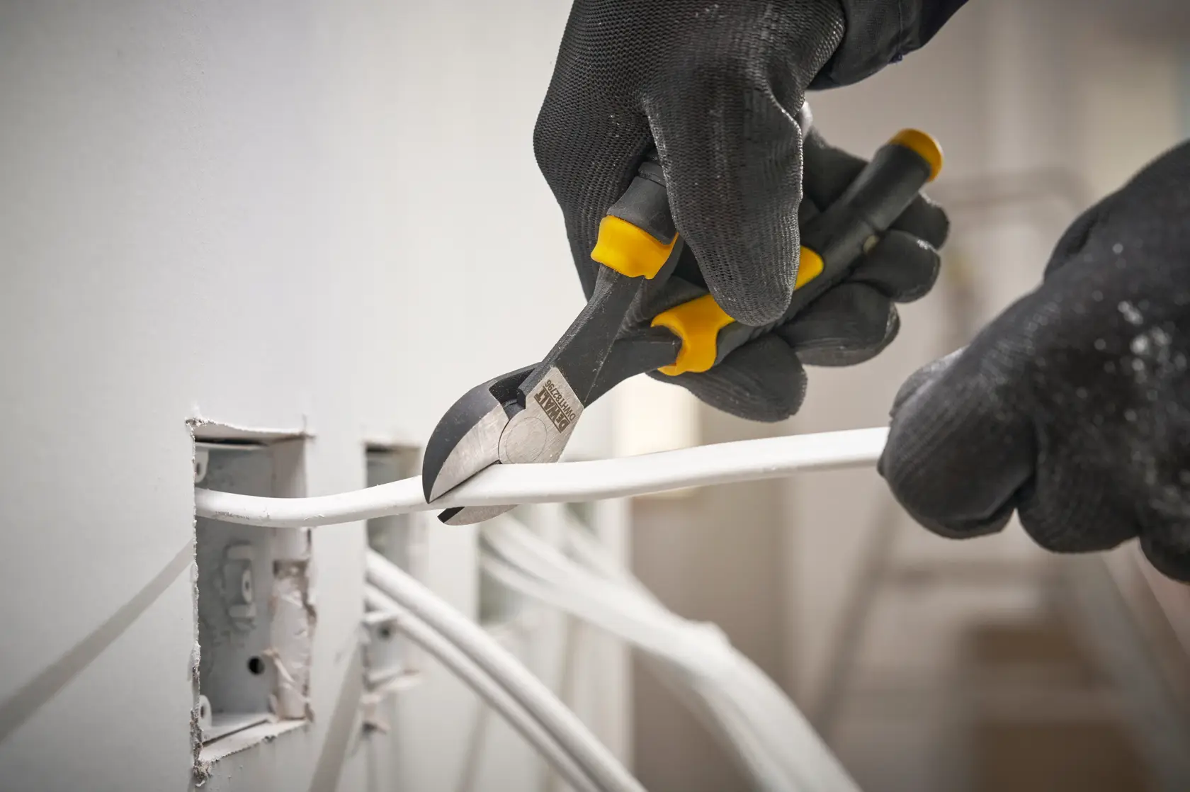 Manos con guantes cortan un cable eléctrico con pinzas DEWALT junto a una toma de pared.