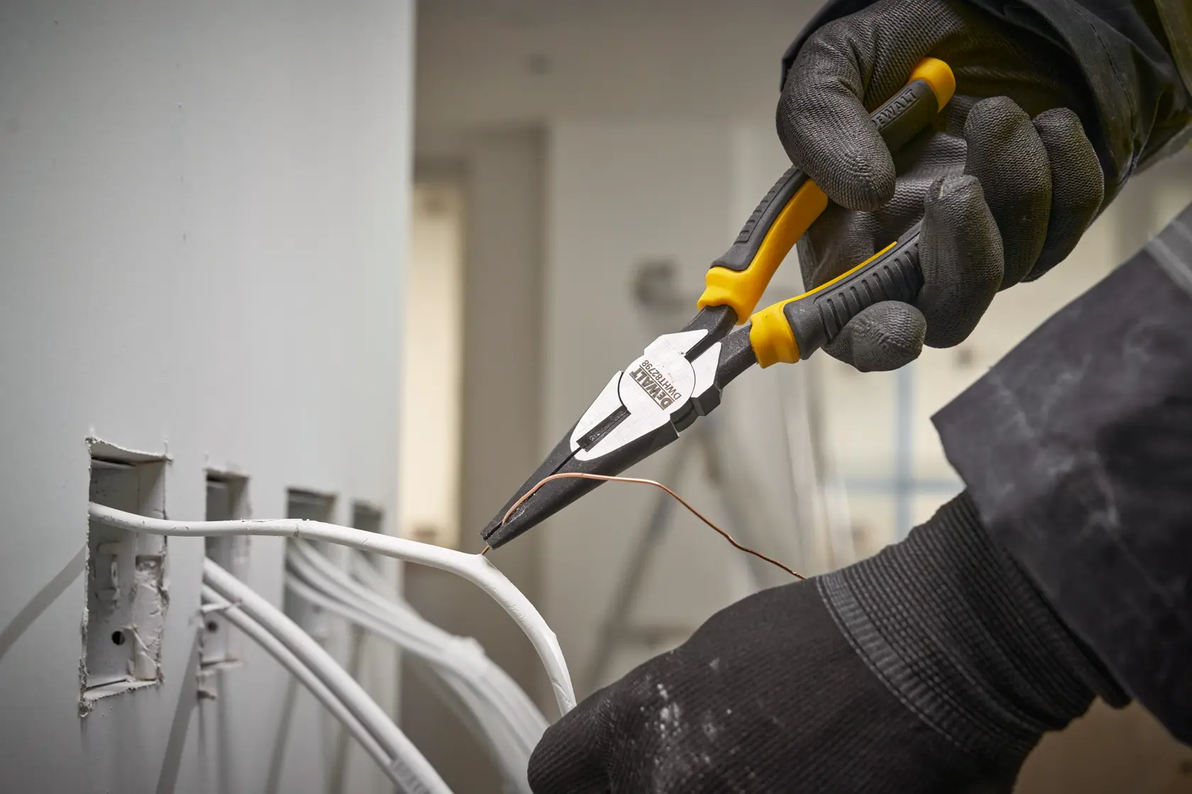 Mano enguantada usa alicates DEWALT para manipular cable cerca de tomas eléctricas en la pared.