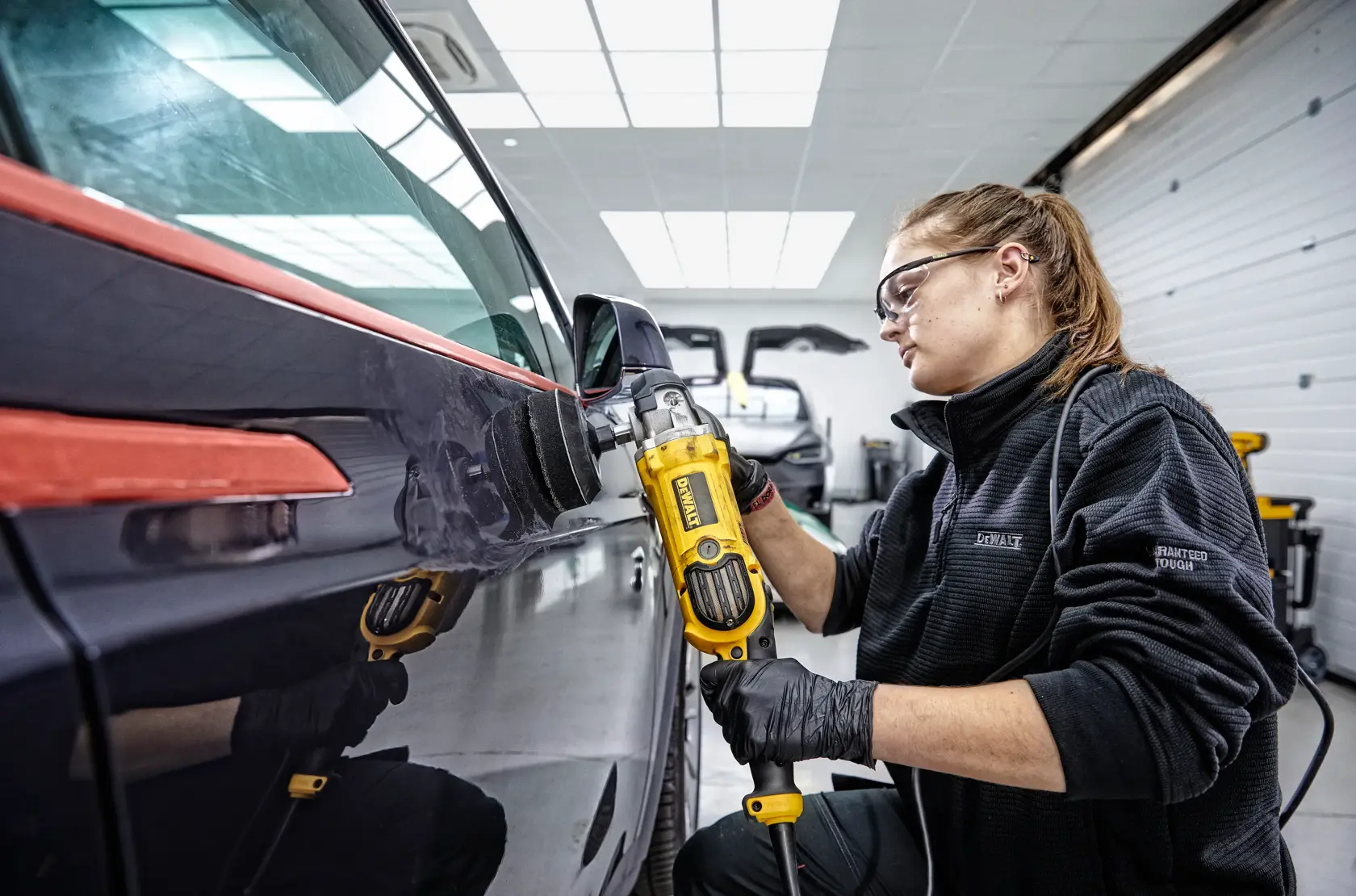 Una persona está usando una pulidora DEWALT DWP849X de color amarillo y negro con una almohadilla de lana grande para pulir sobre un auto negro. La imagen muestra cinta azul en partes del auto y el texto 'PROTECCIÓN E-SWITCH™' en la parte inferior.