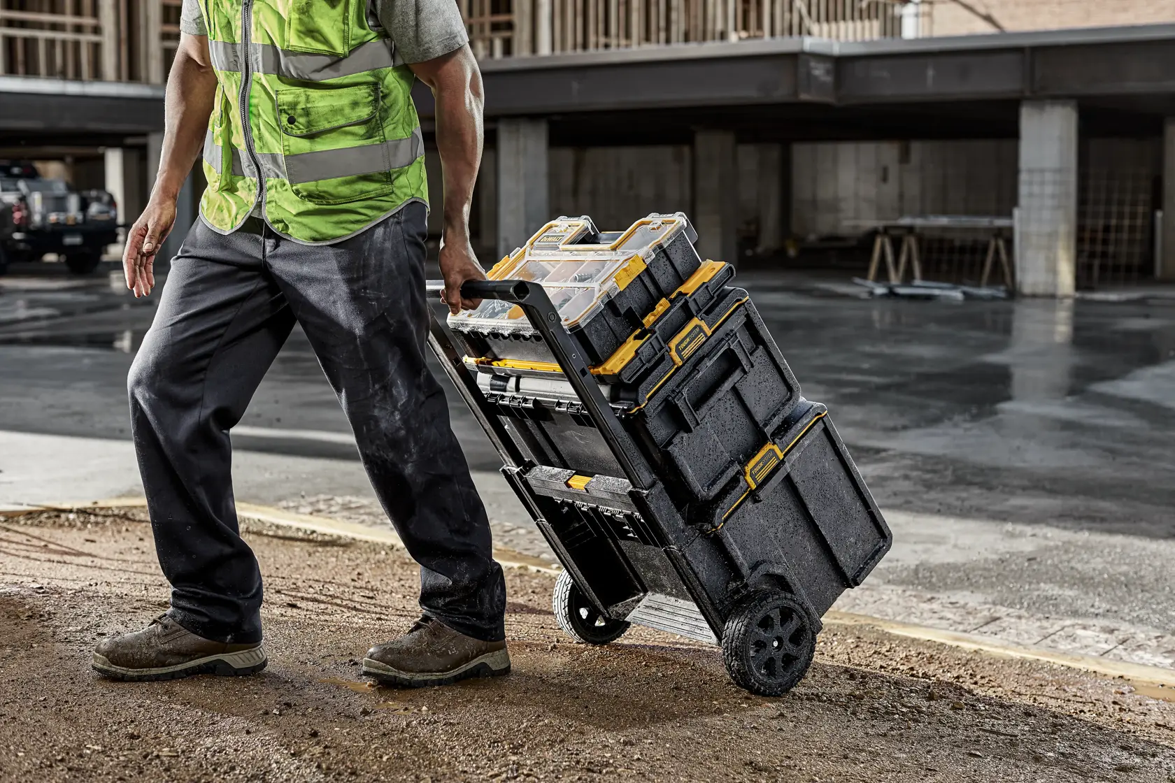 A person wearing a high-visibility vest uses a wheeled cart to transport stacked DEWALT tool boxes on a construction site.