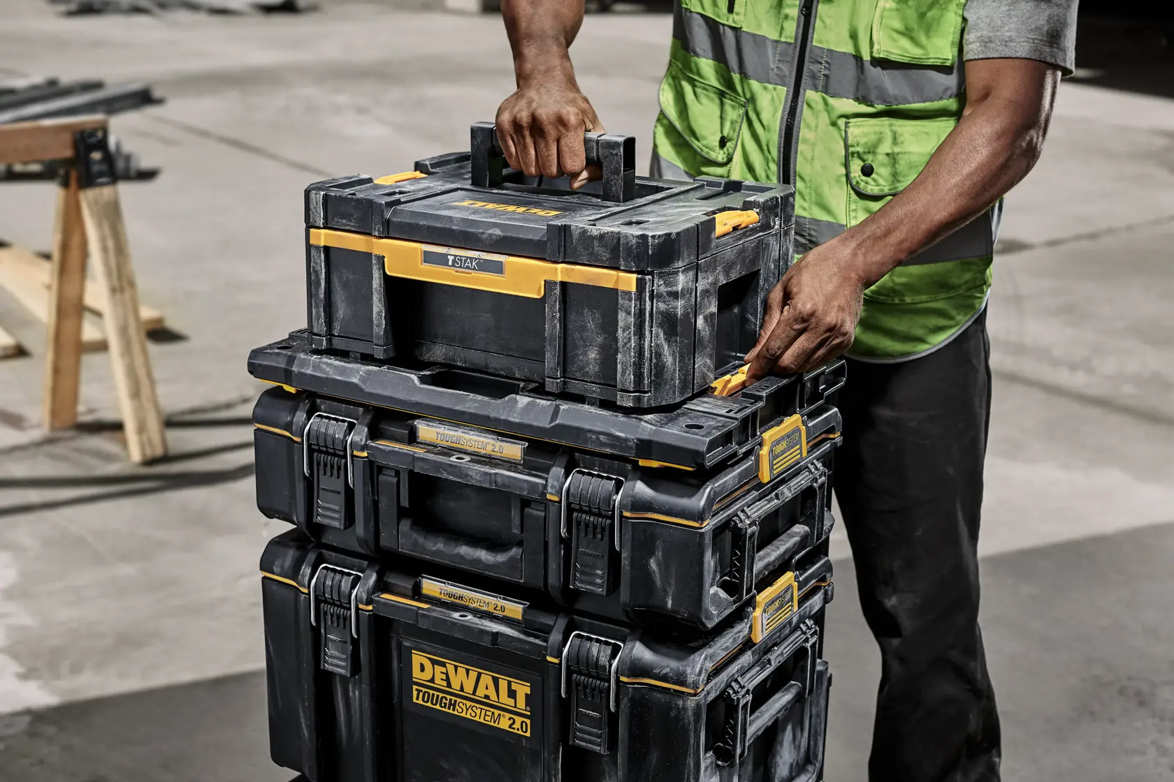 A person wearing a green safety vest is handling a stack of black and yellow DEWALT ToughSystem 2.0 toolboxes outdoors, with focus on the top box being carried.