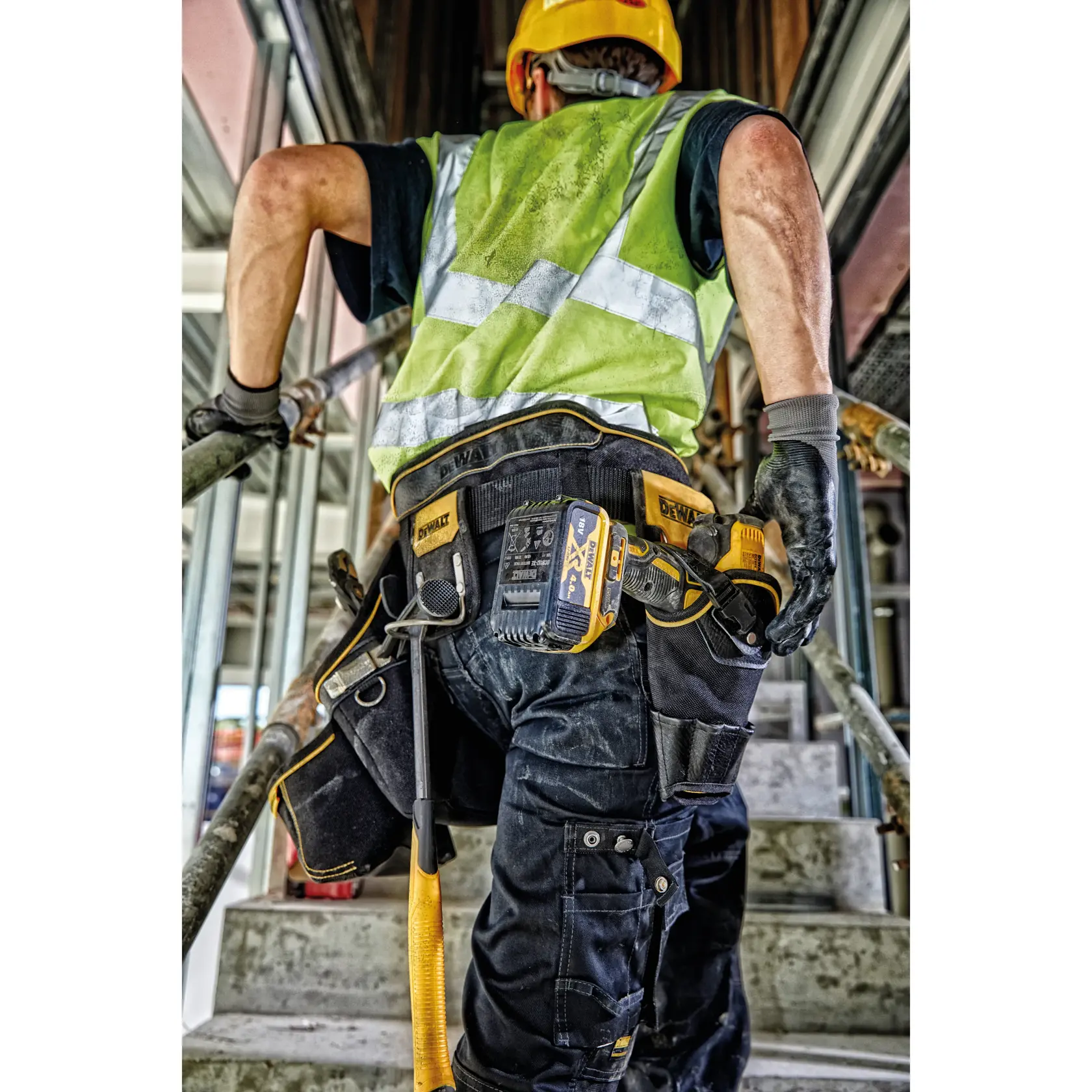 Worker wearing DEWALT tool belt and safety gear climbs stairs on construction site.